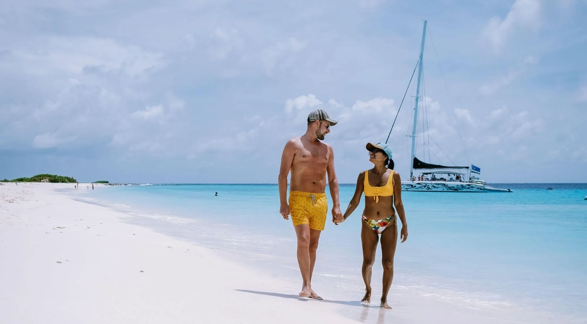 A couple walking Klein Curaçao's empty sand with a Curaçao-flagged catamaran anchored behind them