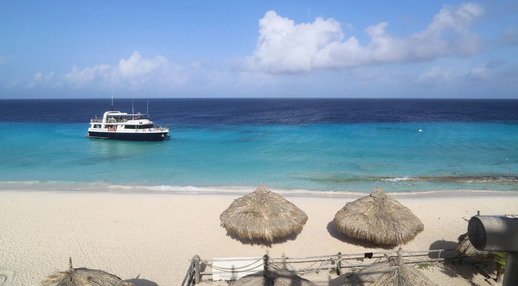 The Mermaid motor yacht anchored off Klein Curaçao's beach with thatched sun shelters in the foreground