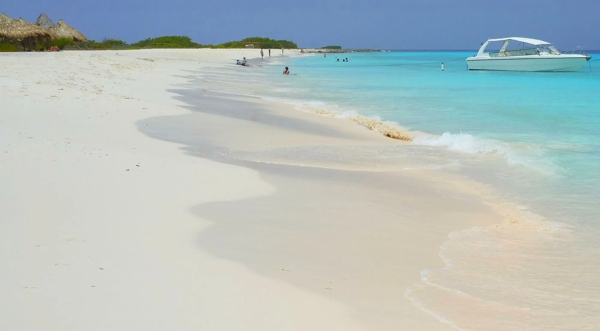 Klein Curaçao's white sand beach with a speedboat moored offshore and thatched palapas along the shore